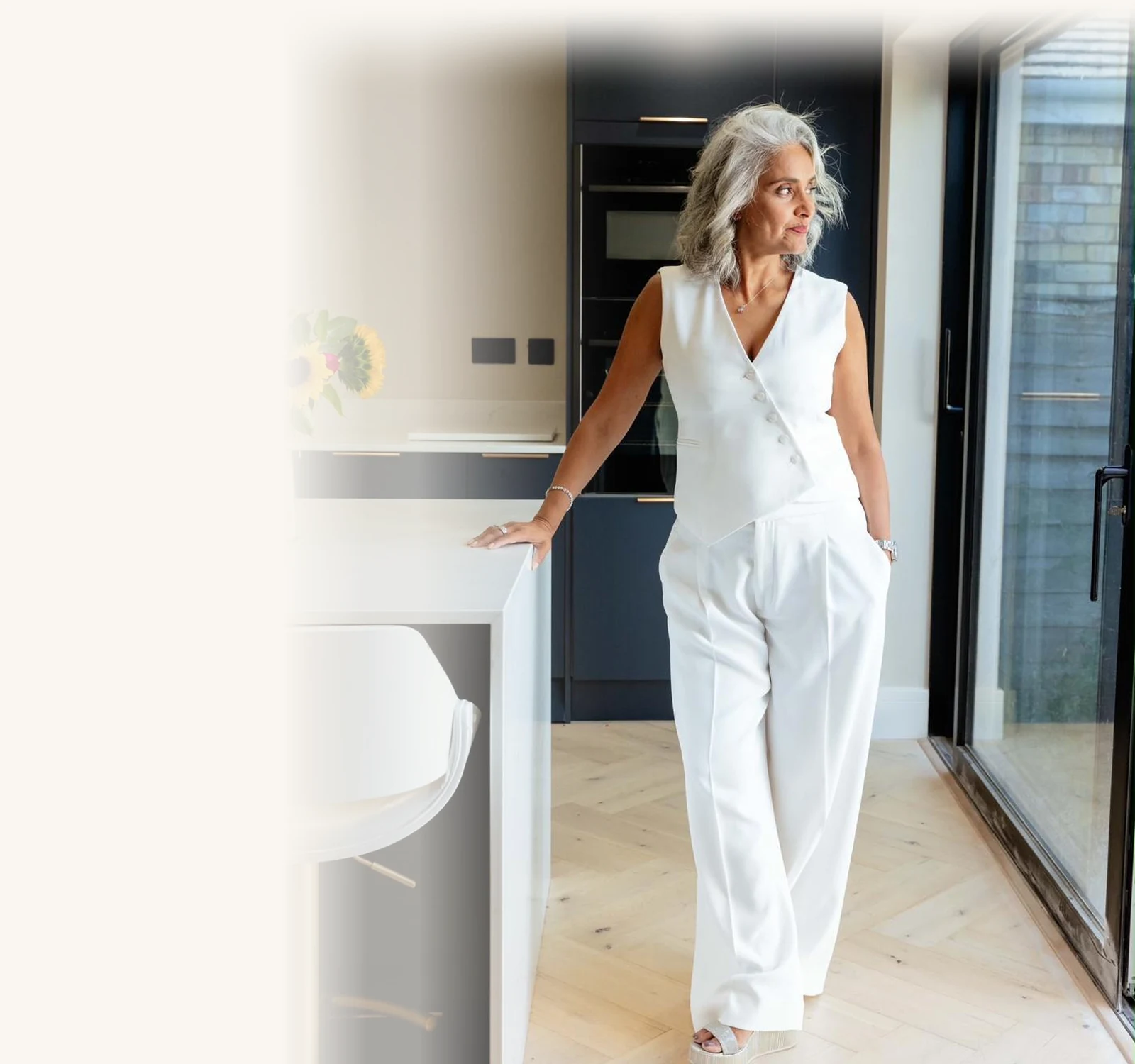 Dimple Amore (Dimple Thakrar) standing in a bright kitchen wearing a white tailored suit, sunflowers on the counter, the portrait from her brand story